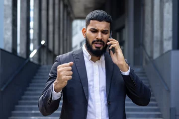 Fotobehang Upset and angry Indian male businessman standing outside an office center and talking on the phone emotionally © Tetiana