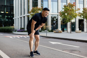 Young Asian male runner standing in the middle of the street bent over, holding his leg and knee,...