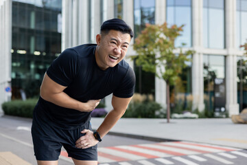 Young Asian male runner standing in the middle of the street bent over, holding his stomach, hiding...