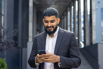 Indian young male businessman standing on the street near an office building and using a mobile phone, dialing a number, writing a message, reading the news - obrazy, fototapety, plakaty