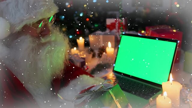 Man in Santa Claus costume at laptop with chromakey screen, holding gift box in hands, among wrapped burning candles, cones, fir branches, garlands, beautiful xmas decoration, close up, background.