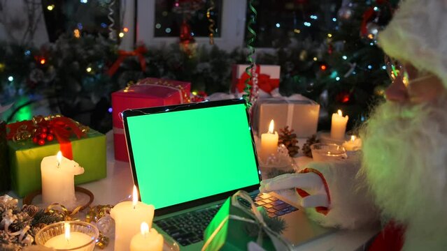 Man in Santa Claus costume sitting at laptop in empty green screen, using keyboard, reading emails with children wishes, next to gifts boxes, candles, fir tree, branches, garlands, close up.