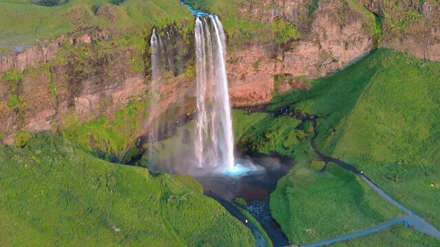Magnificent aerial view of icelandic waterfall falling from a concave cliff flowing out in narrow streams all over meadows with lush greenery on sunny day. High quality 4k footage