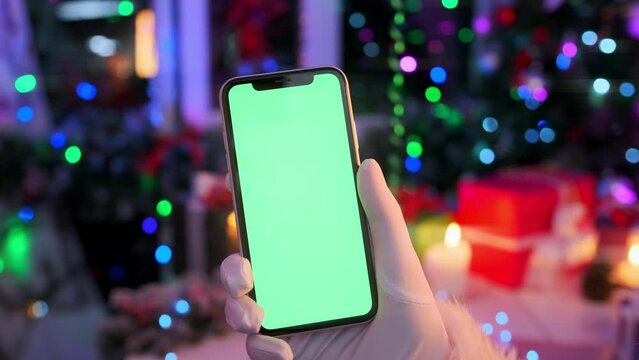 Female hand in white glove holds mobile phone with green chromakey screen against of festive flickering garlands, packaged gift boxes, burning candles and bokeh lights, Christmas background.