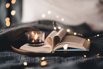 Open paper book with folded pages in heart shape and burning scented burning candle on tray in bed...