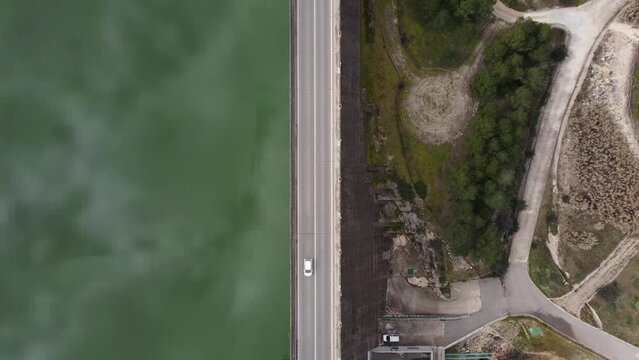 Static drone view from above of a road over a dam wall dividing a reservoir and a wooded area in the center of the image