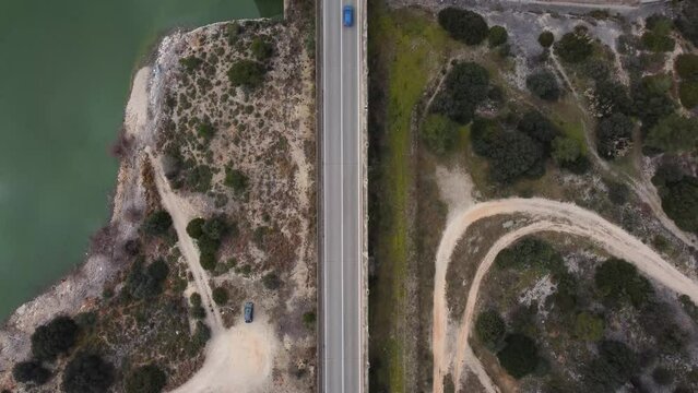 Overhead drone view of a road in a rural area with a lake on one side and a grove of trees on the other with a blue car driving along it