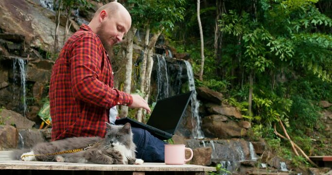 Millennial on jungle pier communicates on laptop with his family and friends on social networks Far from home, man is sad and misses his family. Man caress his fluffy pet and remembers his family