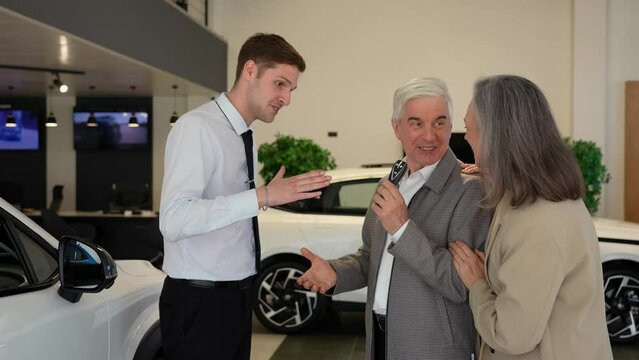 A salesman hands over the keys to a new car to an elderly Caucasian couple.
