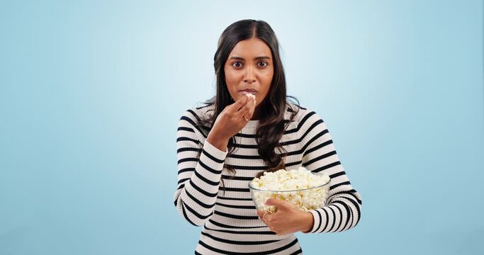 Popcorn, shock and woman in a studio for movie, film or show with entertainment or fun. Surprise, crazy and portrait of young Indian female person eating snack for cinema isolated by blue background.