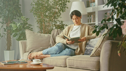 Portrait of beautiful woman relaxing, reading book while relaxing on cozy sofa at home. Smiles,...