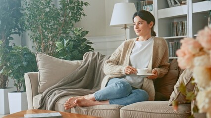 Relaxed woman drinking coffee or herbal tea and feeling carefree and refreshed while relaxing on the couch at home. Break time. Girl enjoying comfortable relaxing sitting on couch in the morning - obrazy, fototapety, plakaty