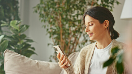 Young beautiful woman sitting on sofa and typing message using smartphone in her apartment. Use...