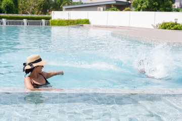 Beautiful Hispanic girl Take dip in the swimming pool happily. while friend jumped into pool until water splashed Woman wearing swimsuit and hat and taking a break from the sun during summer vacation