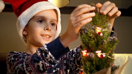 Happy smiling boy gets prepared for Christmas decorating Christmas tree with lights and garlands. Winter holidays, celebrations and party.