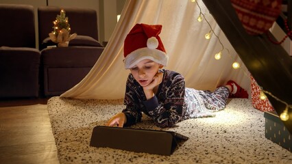 Obraz na płótnie Canvas Little boy in Santa's hat lying in tepee tent and watching video on tablet computer on Christmas eve. Winter holidays, celebrations and party.