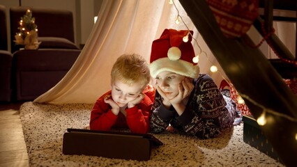 Two boy on Christmas night lying on floor at house and watching video on tablet computer. Winter...