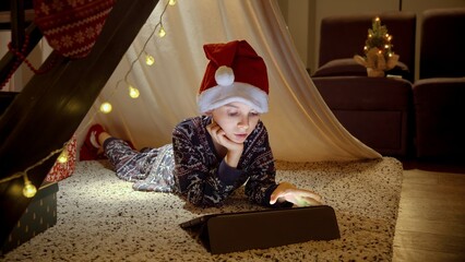 Little boy in pajamas and Santa's hat celebrating Christmas lying in tepee tent and using tablet computer. Winter holidays, celebrations and party.