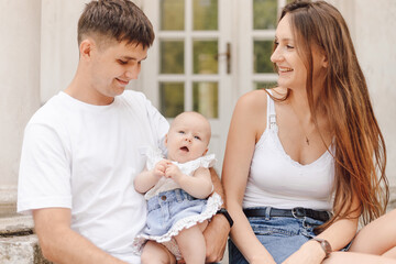 Smiling married couple with little daughter is sitting on summer day against background of house.