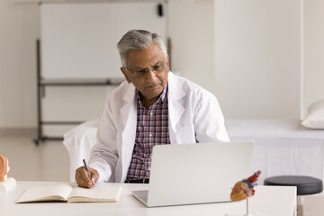 Focused senior Indian doctor man making records in medical history book at laptop, using computer,...
