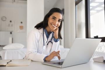 Fototapeta na wymiar Cheerful attractive doctor woman professional portrait. Young Latin practitioner sitting at workplace table with laptop, looking at camera with toothy smile posing for shooting in surgery office