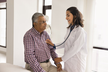 Positive beautiful young doctor examining senior Indian man, touching hand for pulse checking,...