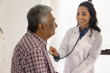 Happy young Latin cardiologist doctor woman listening to heartbeat rate of senior Indian patient,...