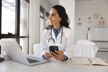 Happy young doctor woman using online medical application on smartphone, holding mobile phone at workplace, giving consultation on Internet, looking away, thinking, enjoying modern technology - obrazy, fototapety, plakaty