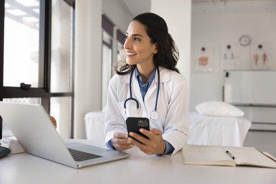 Happy young doctor woman using online medical application on smartphone, holding mobile phone at workplace, giving consultation on Internet, looking away, thinking, enjoying modern technology