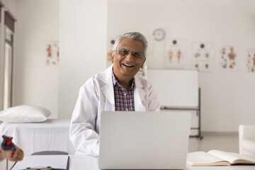 Happy mature senior Indian doctor man wearing white uniform and glasses, sitting at clinic...