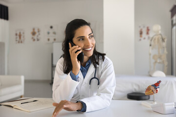 Positive young Latin cardiologist doctor woman talking on mobile phone at workplace, checking...