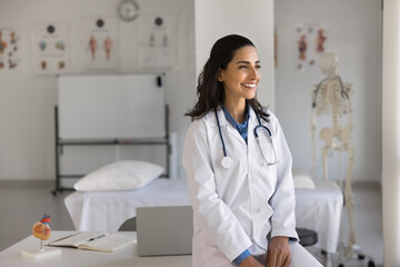 Happy young cardiologist woman posing at workplace, leaning on clinic office table, looking away,...