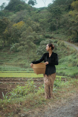 Young woman from an Indonesian village carrying a basket or basket made of woven bamboo. Asian village woman standing and walking in the village rice fields.