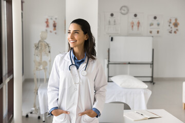 Cheerful beautiful young Latin doctor woman with stethoscope on neck looking at window away with...
