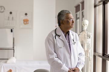 Thoughtful lonely senior Indian doctor standing at clinic window in office, looking away in deep...