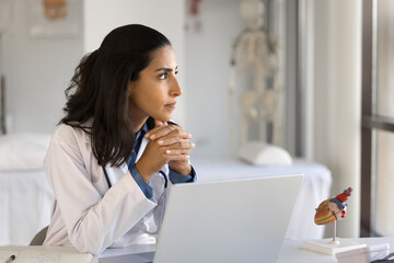 Serious pensive young doctor looking away from workplace, working at computer in office, touching...