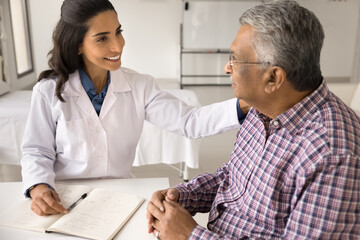 Happy young practitioner woman giving comfort, support to older Indian patient man, touching shoulder, explaining optimistic diagnosis, healthcare examination result, treatment recommendations - Powered by Adobe