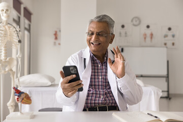 Cheerful senior Indian doctor man giving online consultation to patient on smartphone, waving greeting hand hello at gadget, talking on video call, smiling, laughing