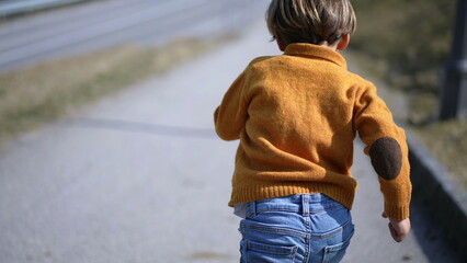 Back of excited small boy sprinting outside during autumn day, Child wearing yellow pullover,...