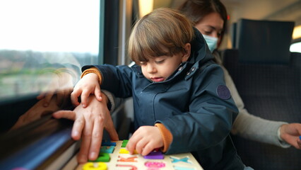 Child seated on mother lap on train ride while doing school lesson activity on the go. Little boy...