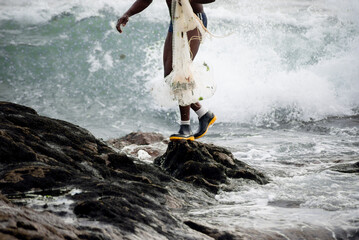 Fisherman walking on top of the rocks holding fishing net. Rio Vermelho beach, city of Salvador, Bahia. - obrazy, fototapety, plakaty