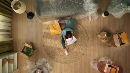 Top view of apartment living room. Woman sitting on the floor reading eviction notice papers, looking upset crying.