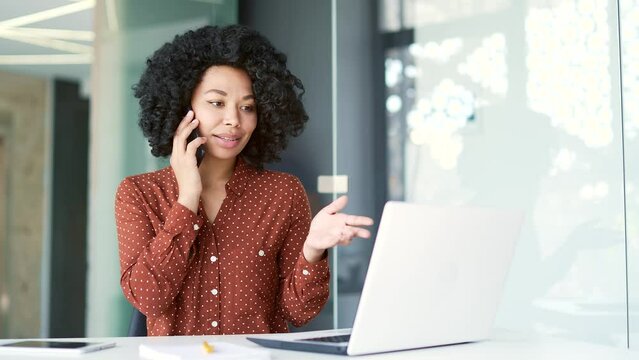 Busy young african american female employee talking on smartphone while sitting at workplace in business office. Focused black woman discusses business with a partner or has a conversation with client
