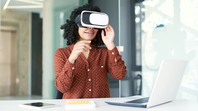 Young african american female employee works using VR glasses in virtual reality simulator while sitting in office. Black woman uses gestures to control computer programs, flips through virtual pages