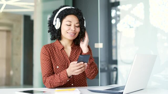 Happy Young African American Female Employee Wearing Headphones Listening To Music Sitting At Workplace In Office. A Smiling Satisfied Black Woman Turns On A Playlist On A Smartphone And Enjoys Music