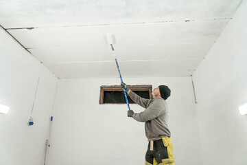 Diligent Worker in Grey Jumper and Yellow Trousers Adds Final Strokes to White Garage Paint Job.