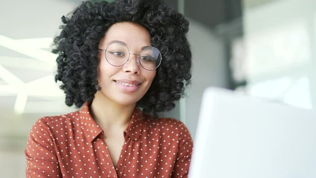 Close up. Young african american female employee typing on laptop sitting at workplace in business office. Black woman in glasses works on a computer on a project, chats online, writes email messages