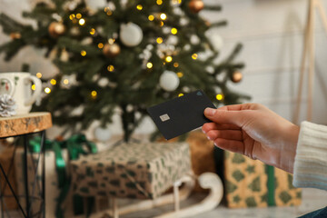 Young woman holding a credit card and a gift box against the background of Christmas decor and gifts, close-up. Christmas and New Year shopping on the Internet, payment by credit card