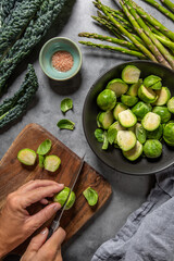 person preparing a healthy dish, top view of a person cutting Brussels sprouts with asparagus and kale on the table, grey background