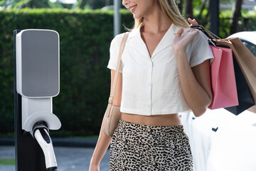 Young woman travel with EV electric car charging in green sustainable city outdoor garden in...